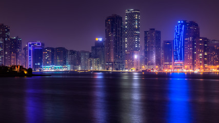 Wall Mural - Beautiful view to Sharjah city center lights skyline at night, United Arab Emirates. Skyscrapers are reflecting in water