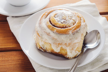 Hot cinnamon bun with sugar creamy icing on white plate on wooden table and napkin. Sweet breakfast or snack with cup of coffee.