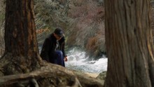 A Rapidly Flowing River Is Framed Nicely Between Trees As A Man Walks Between Them. This Dramatic Slow Motion Shot Is An Interesting Way To Show Man In Nature.