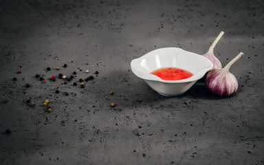 Two garlic next to a white bowl with red sauce. On the stone top lies garlic and gravy, black piper. The concept of eating food, using spices for a meal.