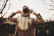 © Strelciuc - Back view of young lady in elegant jacket touching hat while standing on blurred background of autumn countryside.Elegant woman touching hat in nature