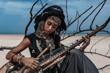 Beautiful young stylish tribal woman in oriental costume playing sitar outdoors