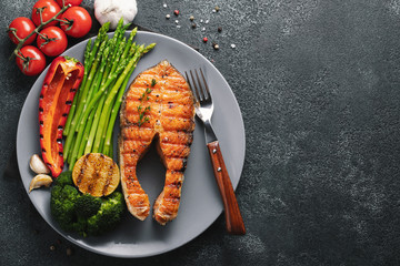 Tasty and healthy salmon steak with asparagus, broccoli and red pepper on a gray plate. Diet food on a dark background with copy space. Top view. Flat lay