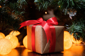 Wall Mural - A gift in craft paper with a red ribbon on the background of a New Year's decorated Christmas tree with lights.