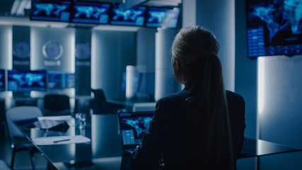 Female Special Agent Works on a Laptop in the Background Special Agent in Charge Talks To Military Man in Monitoring Room. In the Background Busy System Control Center with Monitors Showing Data Flow.