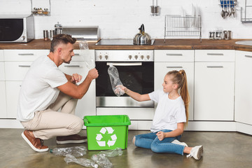 Wall Mural - father and daughter sitting on floor at kitchen and putting empty plastic bottles in box with recycle sign