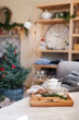 © Marina April - Cozy home interior and decoration, tea time. Wooden serving tray on sofa near Christmas tree with red ornaments. Morning breakfast with mug of hot beverages, white teapot in living room.