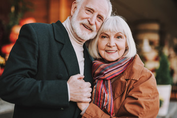 Wall Mural - Portrait of happy bearded gentleman hugging his lovely wife and holding her hand. They looking at camera and smiling