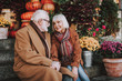 Portrait of bearded gentleman in coat holding hand of his wife while looking at her and smiling. They sitting on stairs with flowers and pumpkins