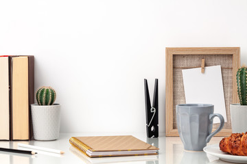 Front view of desk with books, notebook, cactus, mug with coffee, frame and croissant against white wall. Copy space for product display montage...