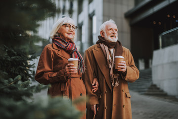 Wall Mural - Portrait of stylish old lady in glasses spending time with husband outdoors. They holding cups of coffee while looking away and smiling