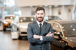 Portrait of a handsome salesman in the suit standing at the showroom with luxury cars on the background