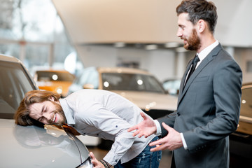 Young man client touching with face car hood of a new luxury car standing with salesperson in the showroom