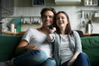 © fizkes - Young millennial joyful couple spend free time together sitting on couch at home. Girlfriend and boyfriend watch on tv favorite show, girl hold remote control switching channels or increases volume