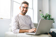 © Louis-Paul Photo - A Young attractive guy is browsing at his laptop, sitting at home on the cozy beige sofa at home, wearing casual outfit