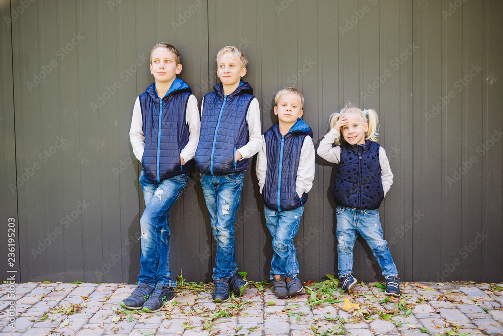 Funny Caucasian big family of three brothers and sister posing standing on growth background of wall in full growth. Equally stylishly dressed in blue vests and jeans. Theme girl in male circle - obrazy, fototapety, plakaty 