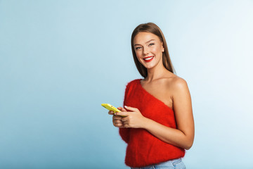 Wall Mural - Smiling young woman posing isolated over blue wall background using mobile phone.