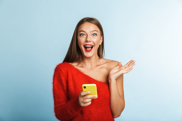 Wall Mural - Emotional excited young woman posing isolated over blue wall background using mobile phone.