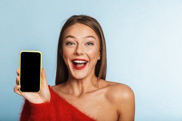 Wall Mural - Excited young woman posing isolated over blue wall background using mobile phone showing empty display.