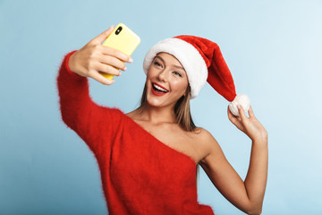 Wall Mural - Excited young woman wearing christmas hat posing isolated over blue wall background take a selfie by mobile phone.