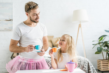 Wall Mural - happy father in pink tutu skirt looking at cute little daughter pretending to have tea party at home