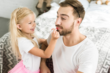 Wall Mural - adorable little daughter applying red lipstick to happy bearded father at home