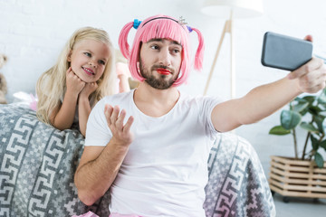 Wall Mural - happy little daughter and father in pink wig taking selfie with smartphone at home