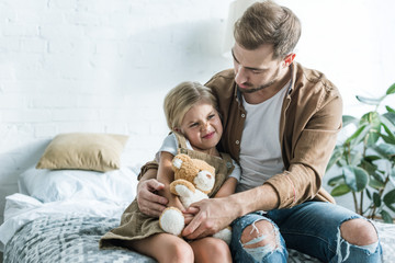 Wall Mural - father looking at little daughter sitting with teddy bear on bed