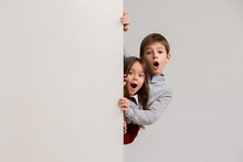 Banner With A Surprised Children Peeking At The Edge With Copyspace. The Portrait Of Cute Little Kids Boy And Girls Looking At Camera Against White Studio Wall. Kids Fashion And Happy Emotions Concept