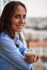 Corporate time-out. Close up portrait of young smiling office woman standing on balcony with cup of coffee. Copy space on right