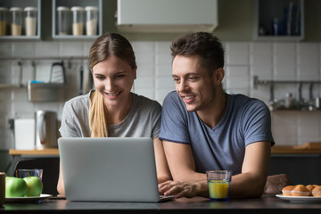 Wall Mural - Happy young smiling couple looking at laptop screen in kitchen together, spending time at home together, using computer for reading online good news, watching video, chatting in social networks