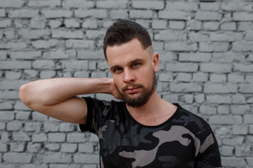 Portrait of attractive young handsome man with a stylish hairstyle and brutal beard in a stylish camouflage military t-shirt near a gray brick wall. Fashionable modern guy.