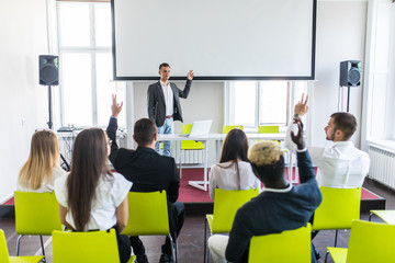 Business coach. Young man gesturing with hand while standing against defocused group of people sitting at the chairs in front of him