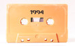 A vintage cassette tape from the 1980s era (obsolete music technology) with the text 1994 printed over it, stencil font. Color: cream, sand. White background.

