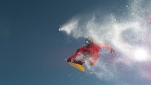 SLOW MOTION CLOSE UP: Snowboarder jumping big air kicker, spraying snowflakes and flying over sun on perfect winter day. Snowboard jump in snow park. Sunbeams shining past jumping boarder in mountains