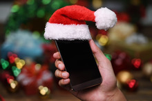 Hand Holding Mobile Phone With Santa Claus' Hat On Top On Christmas Background