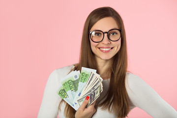 Wall Mural - beautiful young girl in glasses with dollars and euros in her hands on a pink background