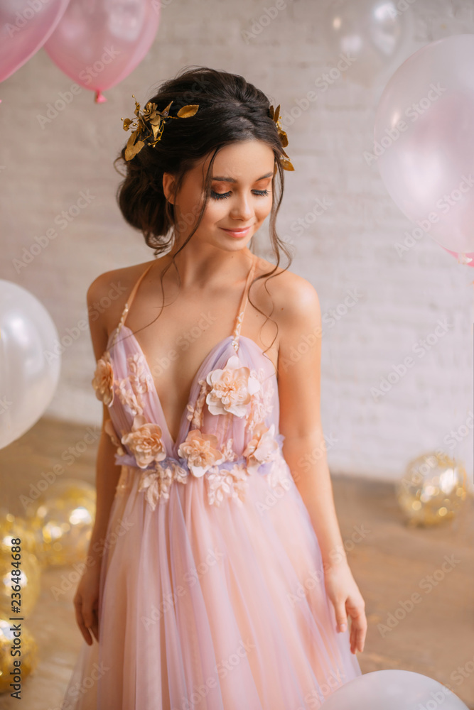 pink dress brown hair A stunning young woman with long, flowing wavy brown hair stands in a soft, natural light setting. She is wearing an elegant strapless dusty rose lace dress with a corset-style bodice and