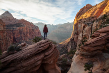 Adventurous Girl At The Edge Of A Cliff Is Looking At A Beautiful Landscape View In The Canyon During A Vibrant Sunset. Taken In Zion National Park, Utah, United States.