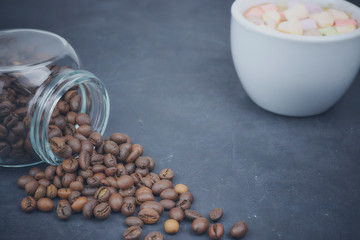 roasted coffee beans poured from a glass jar with a white cup of coffee and marshmallow. angular placement of the coffee object. on dark concrete. horizontal view. copy space.