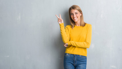 Wall Mural - Beautiful young woman standing over grunge grey wall smiling with happy face winking at the camera doing victory sign. Number two.