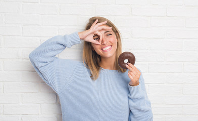 Wall Mural - Beautiful young woman over white brick wall eating chocolate donut with happy face smiling doing ok sign with hand on eye looking through fingers