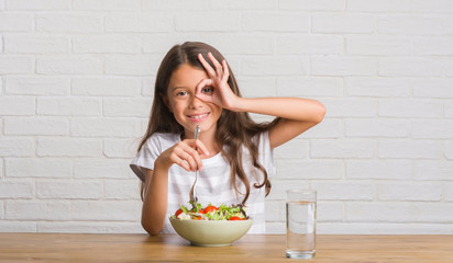 Poster - Young hispanic kid sitting on the table eating healthy salad with happy face smiling doing ok sign with hand on eye looking through fingers