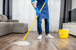 © rh2010 - Man as a professional cleaner in blue uniform washing floor with mopping stick and bucket in the living room of the apartment