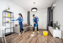 Professional Cleaners In Blue Uniform Washing Floor And Wiping Dust From The Furniture In The Living Room Of The Apartment. Cleaning Service Concept