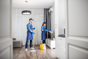 professional cleaners in blue uniform washing floor and wiping dust from the furniture in the living