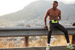 Shot of dark skinned man has intensive preparation to sport competitions, holds bottle of water, volcano in background, looks thoughtfully aside, wears sneakers and leggings, full of energy.