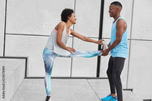 Foto Horizontal view of fit female stretches legs, warms up before race competitons, boyfriend helps her, keeps leg, controls balance, wear sportclothes, look happily at each other