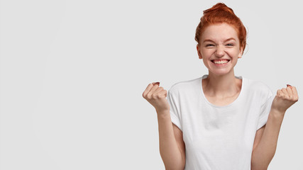 Studio shot of pleasant looking happy girl rejoices from excitement, celebrates excellent news, amused by something positive, raises hands in fists, models over white wall with free space on left