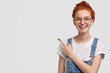 Studio shot of happy freckled lady in glasses, feels satisfied, wears dungarees, indicates at upper left corner, suggests using copy space wisely, stands against white background, has broad smile